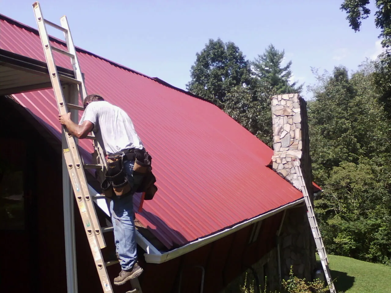 Roofer on a ladder beside a red metal roof and stone chimney — Soffit & Fascia Repair by Roofing Brothers in Rochdale